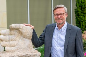 Bürgermeister Otto Steinkamp neben der vor dem Rathaus stehenden Skulptur „Stein des Anstoßes“, an der die Namen der bisherigen Preisträgerinnen und Preisträger zu sehen sind. Foto: André Thöle / Gemeinde Wallenhorst