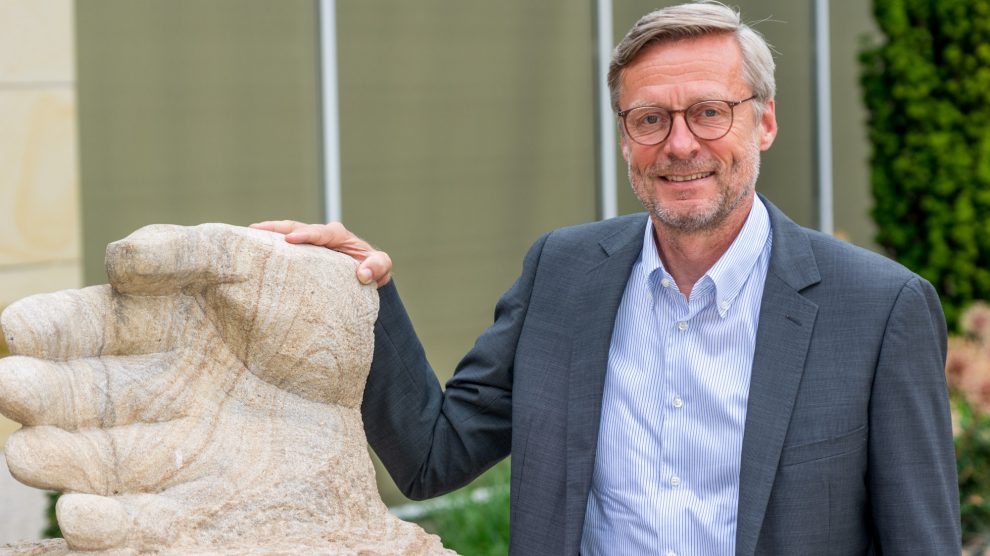 Bürgermeister Otto Steinkamp neben der vor dem Rathaus stehenden Skulptur „Stein des Anstoßes“, an der die Namen der bisherigen Preisträgerinnen und Preisträger zu sehen sind. Foto: André Thöle / Gemeinde Wallenhorst