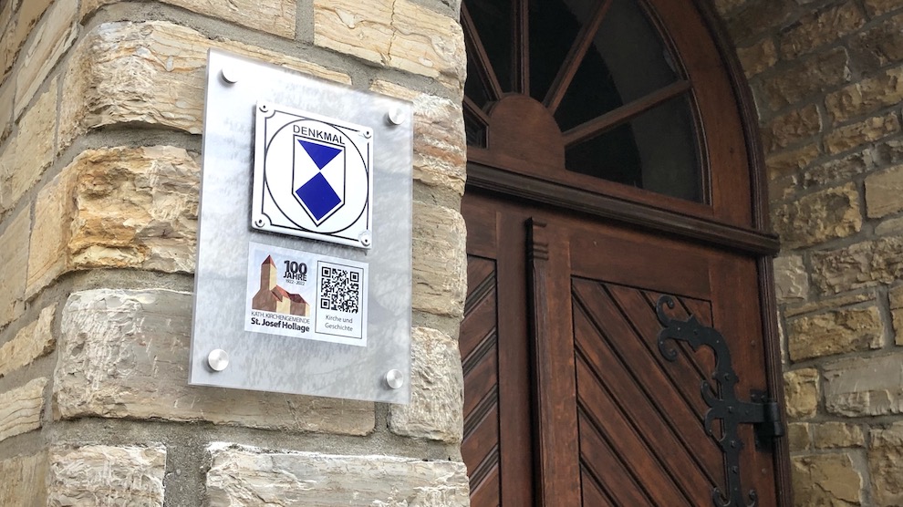 Ein neues Hinweisschild am Haupteingang der Kirche St. Josef Hollage weist ab sofort auf das Jubiläum anlässlich des 100-jährigen Bestehens der Kirchengemeinde hin. Foto: Volker Holtmeyer